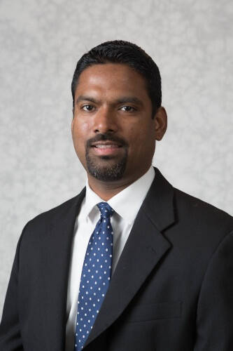 A man looks into a  camera. He is wearing a black suit, white shirt and blue and white polka dot tie.